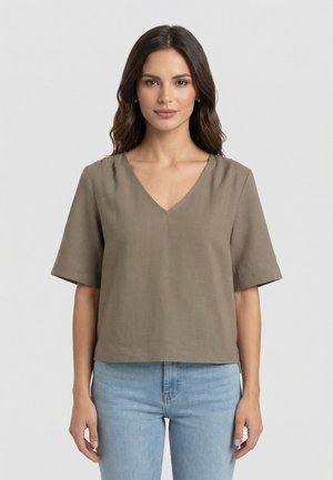 Femme aux cheveux bruns mi-longs portant un haut taupe à manches courtes et col en V, ainsi qu'un jean bleu clair, debout devant un fond uni.