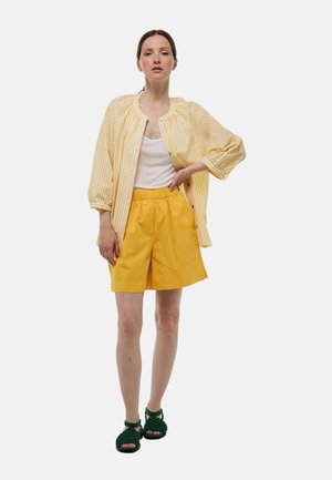 Femme debout portant une chemise oversize rayée jaune, un débardeur blanc, un short jaune et des sandales vert foncé sur un fond uni.