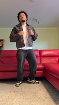 Crna i siva jakna sa uzorkom, bijela majica, crne traperice i crne Nike tenisice. Stoji pored crvene kožne sofe, u zatvorenom.