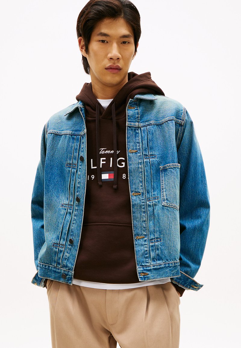 Τζιν  μπουφάν σε ανοιχτό μπλε με κλασικό σχεδιασμό, που διαθέτει μπροστινή τσέπη, συνδυασμένο με καφέ φούτερ με κουκούλα που φέρει λογότυπο, ελαφρύ πουκάμισο από κάτω.