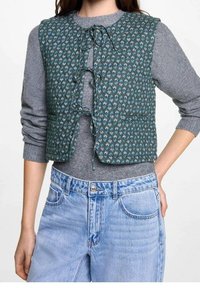 Tkanina w kolorze turkusowym, pikowany kamizelka z kwiatowym wzorem, z zapięciem na wiązanie z przodu, w połączeniu z szarym długim rękawem i jasnoniebieskimi jeansami.