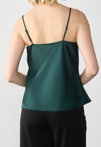 Camisole in raso verde scuro con spalline regolabili, texture liscia e vestibilità rilassata. L'orlo scende sotto la vita.