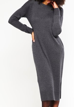Vrouw draagt een lange, donkergrijze, gebreide sweaterjurk met lange mouwen en een zijsplit, poserend met één hand op haar heup en de andere bij haar gezicht.