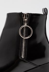 Bottine en cuir verni noir avec une finition brillante, une fermeture éclair proéminente avec une tirette circulaire, et des détails cousus le long de la couture.