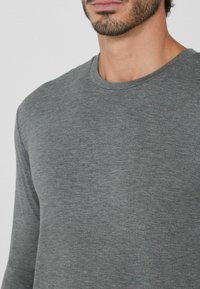 Mann mit kurzem Bart, der ein einfaches graues Langarmshirt mit Rundhalsausschnitt trägt, von Hals bis zur Mitte des Oberkörpers vor einem weißen Hintergrund gezeigt.