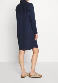 Hemd Kleid in Marineblau mit langen Ärmeln, Knopfmanschetten und lockerer Passform. Hergestellt aus glattem Material, mit einem Rückenpasse-Design und seitlichen Schlitzen.