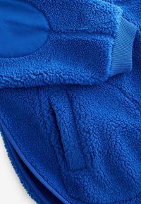 Veste en polaire bleue avec un tissu à poils doux et texturé, des poignets côtelés et une poche avant avec une ouverture arrondie.