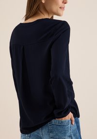 Blouse à manches longues bleu marine avec une texture côtelée, un col rond et un dos plissé. Portée avec un jean bleu clair, mains dans les poches.