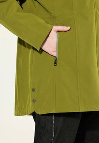 Giacca verde oliva con una texture liscia, tasca laterale con zip e dettagli con bottoni a pressione all'orlo. Vestibilità sartoriale, design contemporaneo.