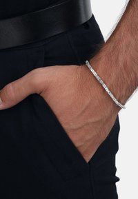 Silvervävd armband som visas på ett handled, med ett strukturerat mönster. Handen är delvis dold i en mörk ficka, med ett svart bälte synligt.