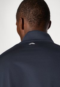 T-shirt polo azul-escuro com colarinho e um pequeno logotipo branco nas costas, feito de um tecido liso e respirável. Cabelo texturizado visível.