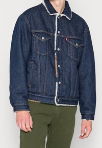 Chaqueta de mezclilla en azul oscuro con cuello de borreguito. Presenta dos bolsillos en el pecho y botones de metal. Dobladillo desgastado y forro interior visible.