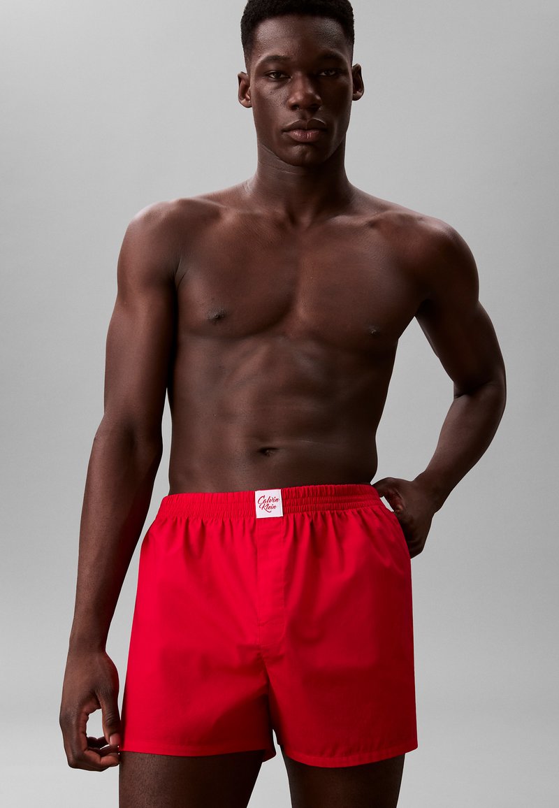 Pantaloni corti rossi realizzati in tessuto liscio, con un elastico in vita e un'etichetta bianca con il logo "Calvin Klein". La vestibilità è morbida e informale.