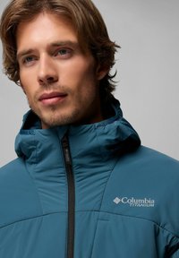 Teal wasserfeste Jacke mit hohem Kragen, schwarzem Reißverschluss und Columbia Titanium-Logo. Sie hat eine glatte Textur und ein gepolstertes Design.
