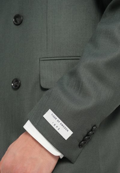 Blazer verde scuro realizzato in tessuto texture. Presenta un'etichetta bianca con "TIGER OF SWEDEN 1903" e tre bottoni neri sul polsino.