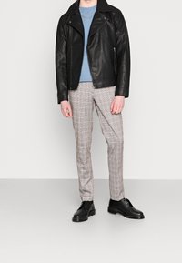 Černá kožená motorkářská bunda, modré tričko s kulatým výstřihem, světle hnědé kostkované kalhoty a černé boty v postoji na neutrálním pozadí.