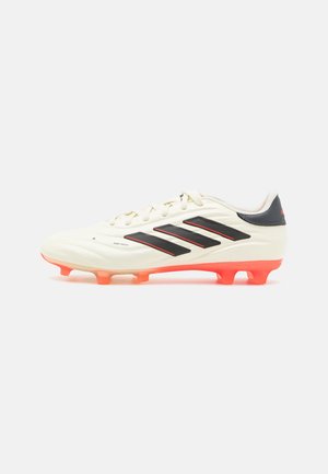 Botte de football blanche avec des rayures noires et des accents rouges. Présente un matériau lisse avec une surface texturée et des crampons oranges.