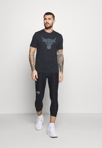 Sort T-shirt med okse grafisk design, sorte kompressions leggings og hvide træningssko. Bemærkelsesværdige tatoveringer på armene. Enkel design, tætsiddende stil.