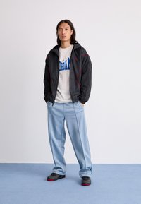 Person med langt hår iført en sort jakke, hvid Jordan-logo t-shirt, lyseblå Carhartt bukser, og sorte sneakers står på en blå gulv.