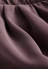 Material moale, drapat într-o nuanță subtilă de mauve, cu o textură netedă și un luciu fin, prezentând detalii adunate de-a lungul marginii superioare.