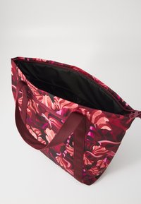 Un grand sac fourre-tout avec un motif floral dans des nuances de rouge et de rose, doté de poignées solides et d'un dessus non zippé dévoilant un intérieur noir.
