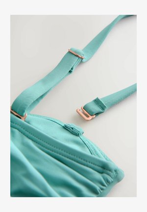 Tyrkysová podprsenka s nastaviteľnými ramienkami, vybavená detailom z ružovo-zlatého kovania. Hladká textúra s švom a zapínaním na háčiky.