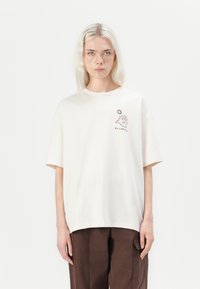 White cotton T-shirt with short sleeves, featuring a brown embroidered design of a face and sun, paired with brown pants.