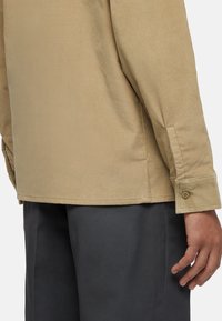 Beige corduroy blouse met lange mouwen, met knoopmanchetten en een rechte zoom, gedragen over donkere broek. Soepele textuur en subtiele ribbels.
