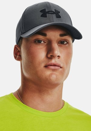 Gray cap with a black Under Armour logo, flat brim, and structured design. Worn with a bright green t-shirt and short hair.