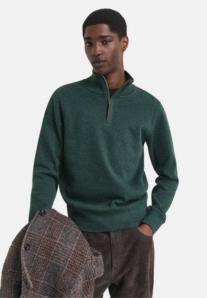 Sweater verde de media cremallera en tejido suave con puños acanalados, combinado con un abrigo de lana de cuadros marrón que el modelo sostiene bajo el brazo.