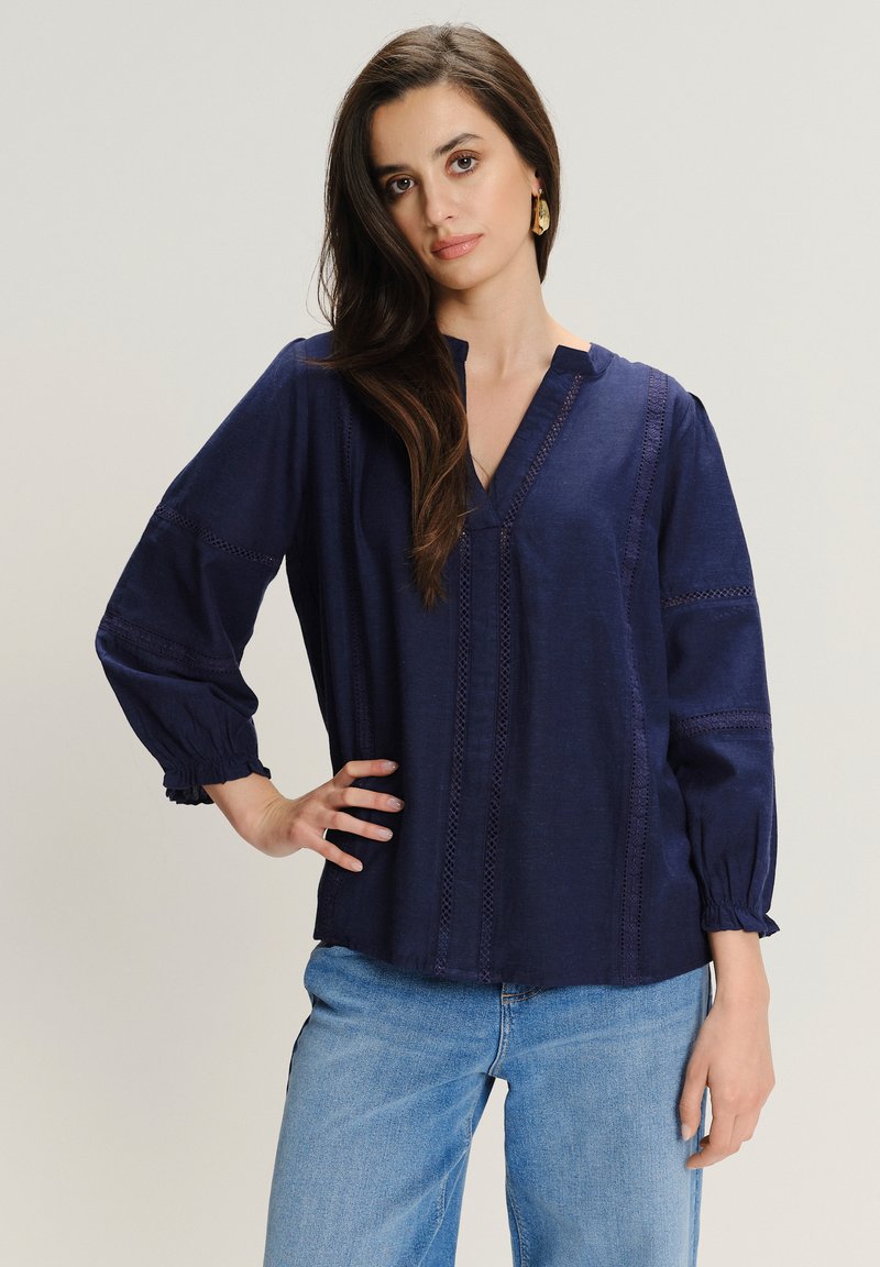 Granatowa lniana bluzka z długimi, bufkami, dekoltem w kształcie litery V oraz ozdobnym przeszyciem wzdłuż przodu i rękawów. Luźny krój.
