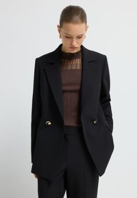 Czarna marynarka o strukturalnym fasonie, z złotymi guzikami akcentującymi i ostrymi klapami, noszona na brązowej bluzce z prążkowanego materiału z przezroczystymi detalami.