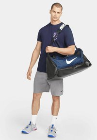 Bolsa de gimnasio de la Marina con fondo de cuero negro, gran logo blanco de Nike, correa ajustable y asas dobles. Modelo en atuendo deportivo, pantalones cortos grises y zapatillas.