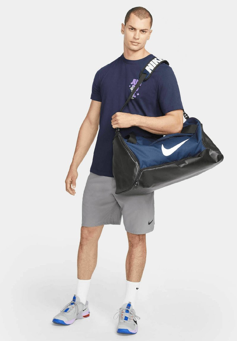 Bolsa de gimnasio de la Marina con fondo de cuero negro, gran logo blanco de Nike, correa ajustable y asas dobles. Modelo en atuendo deportivo, pantalones cortos grises y zapatillas.