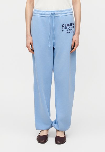 Personne portant un pantalon de survêtement ample bleu clair avec un texte noir sur la cuisse droite et des chaussures marron foncé, debout sur un fond blanc.