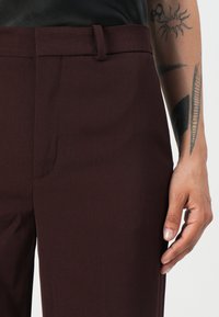Bordeauxfarbene maßgeschneiderte Hose mit glatter Textur, ausgestattet mit einem Bund und einer seitlichen Tasche. Eine sichtbare Hand ruht auf der Hose.