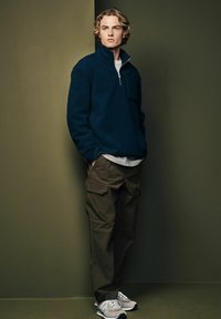Pull en polaire navy avec col haut et fermeture éclair ; pantalon cargo olive avec poches ; baskets grises avec accents clairs. Texture, coupe décontractée.