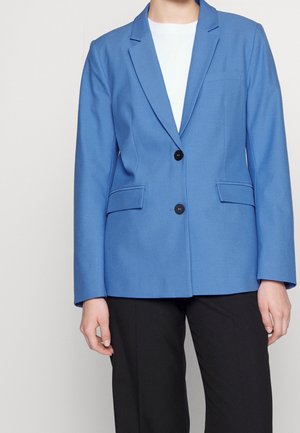 Blå blazer med skräddarsydd passform, två svarta knappar, sidfickor och notch-kragar. Buren över en vit topp, matchad med svarta byxor.