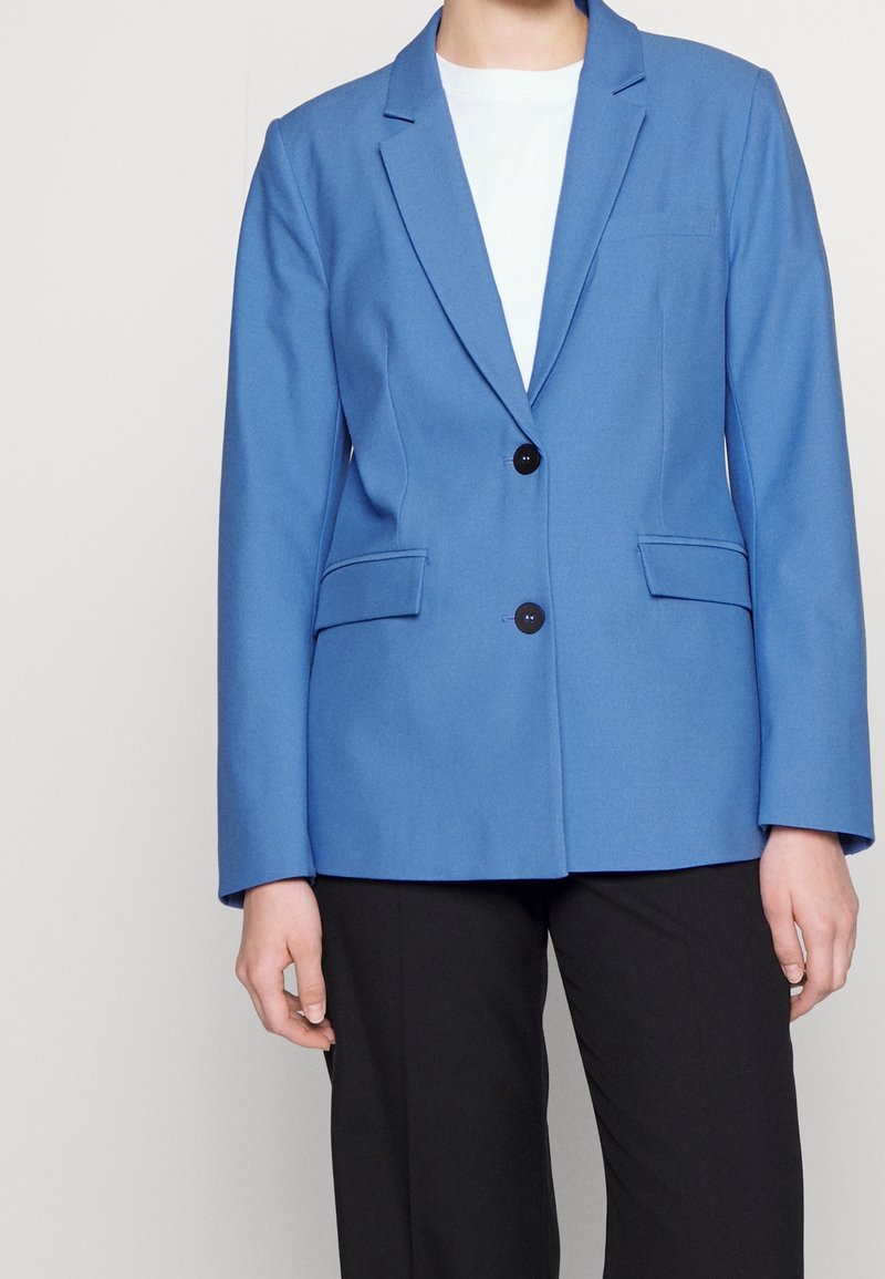 Niebieski żakiet o dopasowanym kroju, z dwoma czarnymi guzikami, bocznymi kieszeniami i klapami typu notch. Noszony na białej bluzce, w połączeniu z czarnymi spodniami.