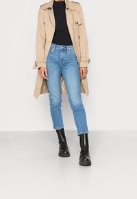 Persona con abrigo trinchera beige, top negro de canalé, jeans azul claro y botas de tobillo negras contra un fondo blanco liso.