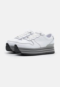 Pár bílých platformových tenisek se silnými šedými pruhovanými podrážkami, bílými šňůrkami a stříbrným detaily.