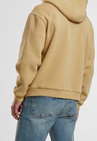 Beiger Baumwoll-Hoodie mit Kapuze, gerippten Bündchen und Saum. Kombiniert mit hellblauen Jeans aus Denim mit Gesäßtaschen. Glatte Textur.