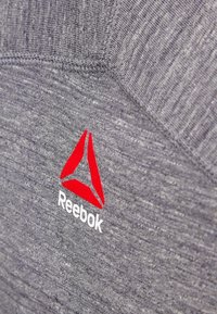 Tessuto atletico grigio con una texture mélange, caratterizzato da un logo Reebok rosso e accenti geometrici bianchi. Ripresa ravvicinata che mette in evidenza i dettagli.