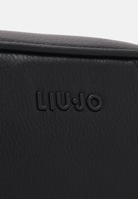 Dettaglio di una borsa a tracolla in pelle nera con logo "LIU·JO" impresso su una superficie testurizzata. Forma rettangolare con linee pulite e design minimale.