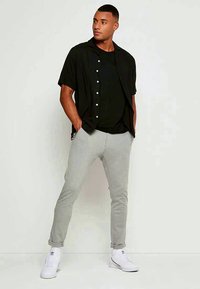 Must tee-nup kätikutsega lühikesed varrukad musta särk musta T-särgi peal, hallid istuvad dressipüksid ja valged tossud. Neutraalne taust.