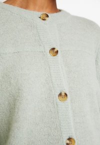 Cardigan vert menthe clair en tissu doux et texturé, avec de gros boutons marron et un col rond bordé de côtes.