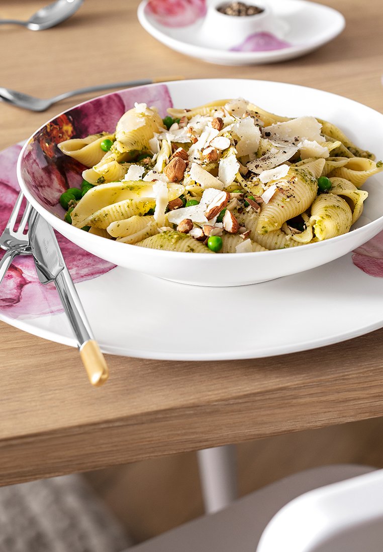 Biała miska z zielonym makaronem i migdałami na talerzu z różowymi akcentami; tło drewnianego stołu; widelec i nóż ułożone obok dania.