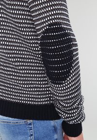 Pull tricoté avec des rayures noires, blanches et brunes ; il présente des coudières texturées et des poignets côtelés. Coupe décontractée, matériau doux.