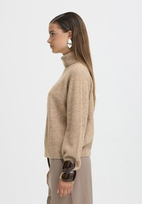 Maglione beige a collo alto con maniche lunghe. Tessuto morbido, vestibilità rilassata. Accento marrone sulla manica con tre sezioni arrotondate.