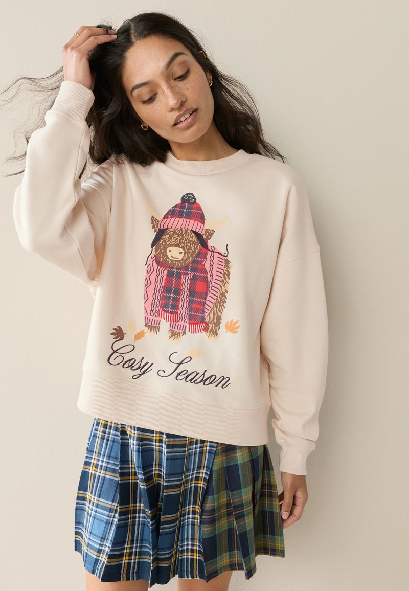 Sudadera de color rosa claro con un gráfico de un toro de dibujos animados que lleva un sombrero y bufanda a rayas, combinada con una falda de cuadros azul y verde.